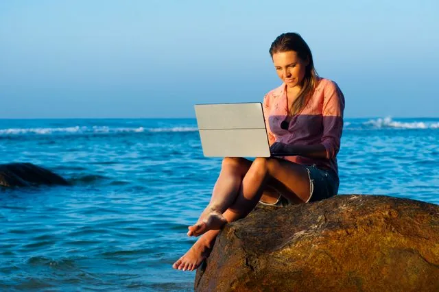 Vacaciones y ansiedad: por qué nos cuesta tanto dejar de ser productivos 3 Mujer trabajando con el portátil sentada en una roca junto al mar, ejemplo de hiperconectividad en vacaciones